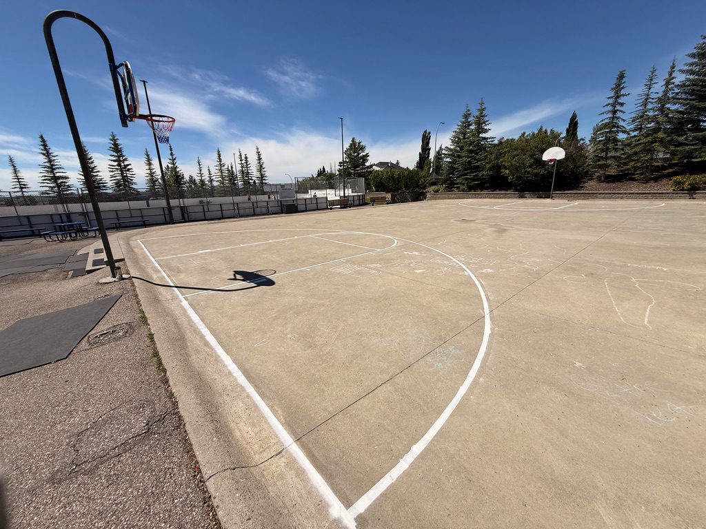 The Signal Hill outdoor basketball court on a bright summer day, two hoops on a painted concrete pad beside the rink boards, evergreens and blue sky behind.